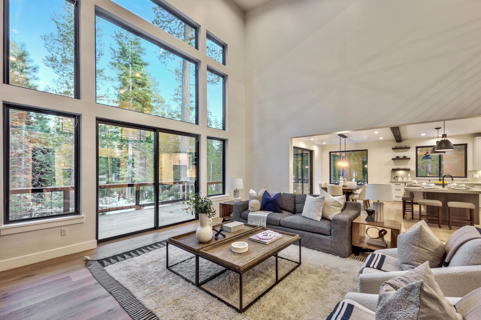 Great room with floor-to-ceiling windows framing tall pines, open to the kitchen beyond.