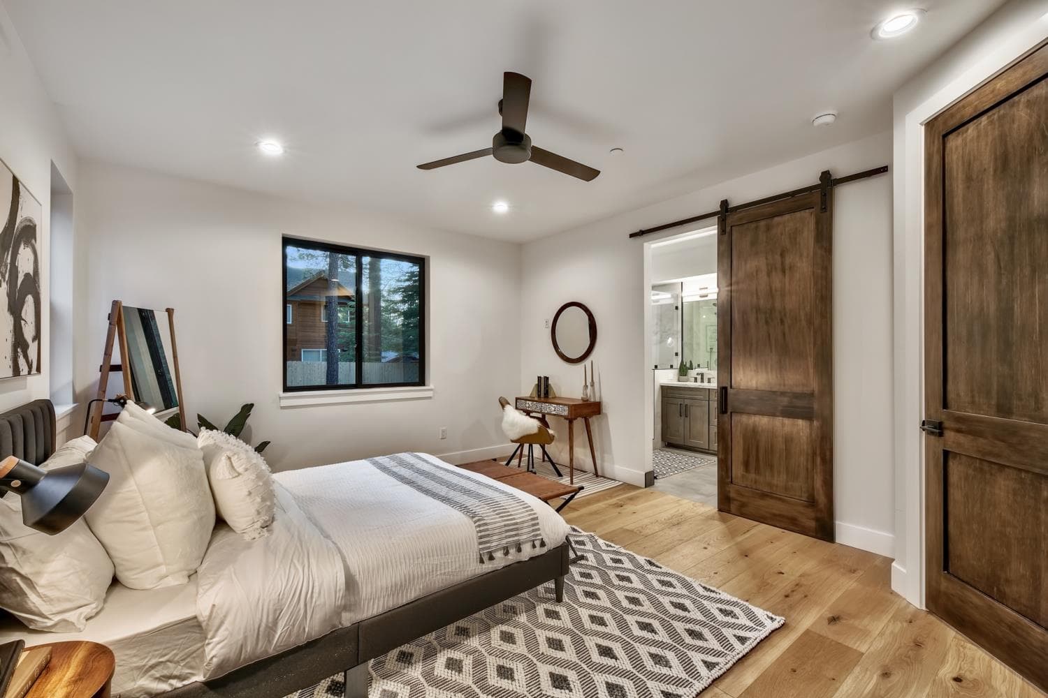 Primary bedroom with wide-plank wood floors, a barn door to the ensuite, and forest views.